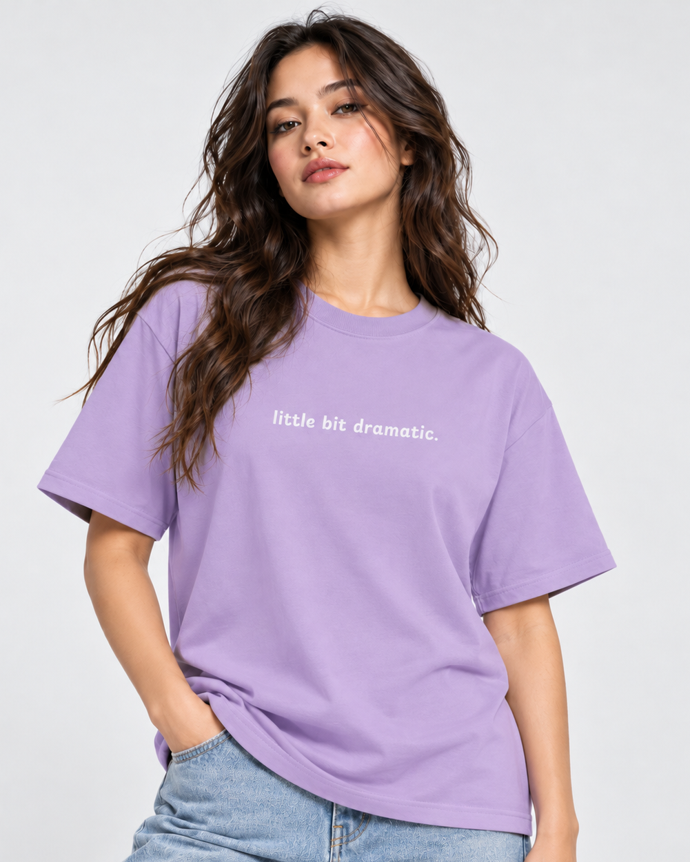 A woman wearing a lavender purple short-sleeve t-shirt with the text 'little bit dramatic.' printed in white across the chest, paired with light blue jeans, against a plain light gray background.