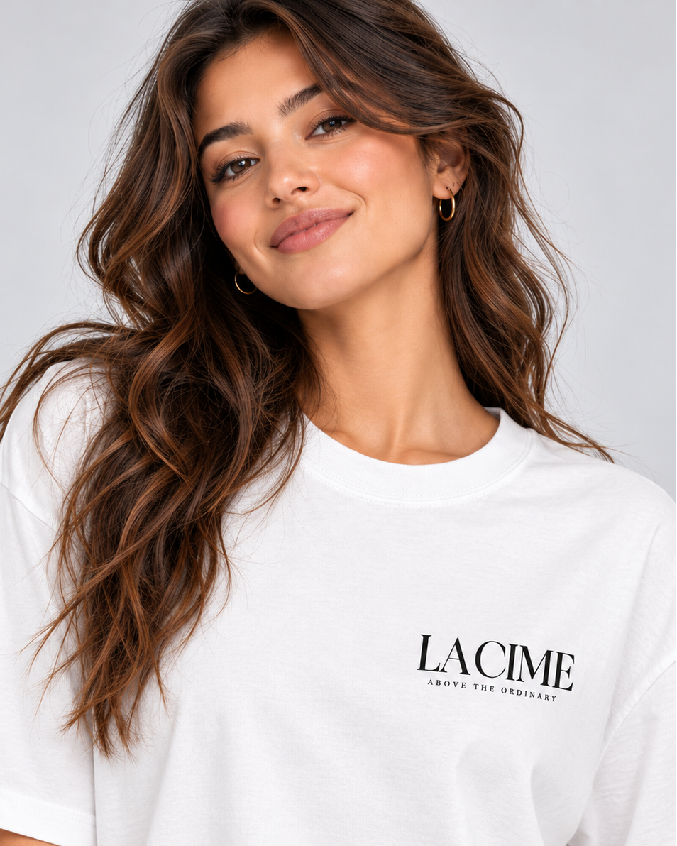 A woman with long wavy brown hair wearing a white oversized Lacime t-shirt with black text on the left chest, photographed against a light gray studio background.