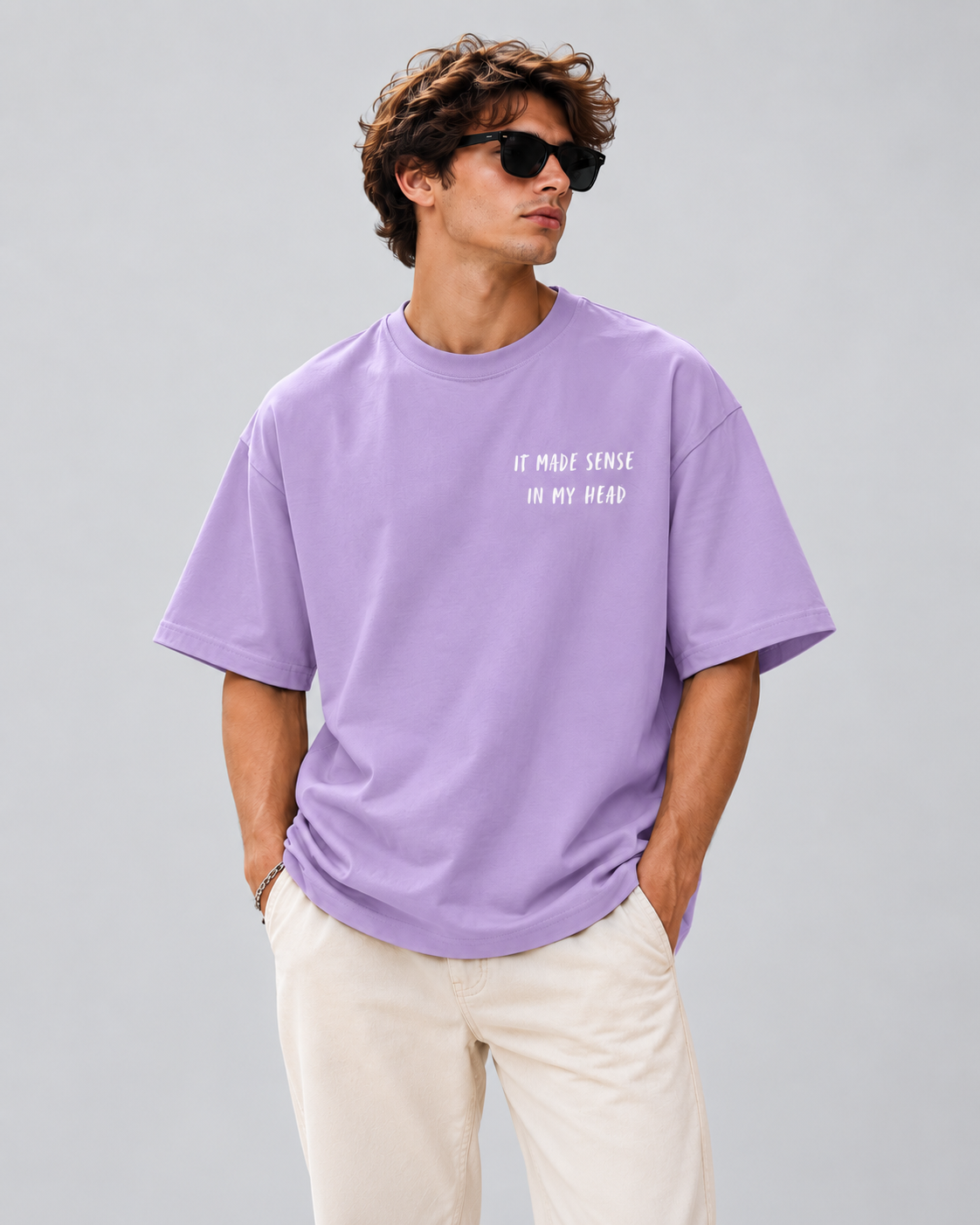 A man wearing a lavender purple oversized t-shirt with the white printed text 'IT MADE SENSE ON MY DEAD' and cream-colored pants, standing against a solid gray studio background.